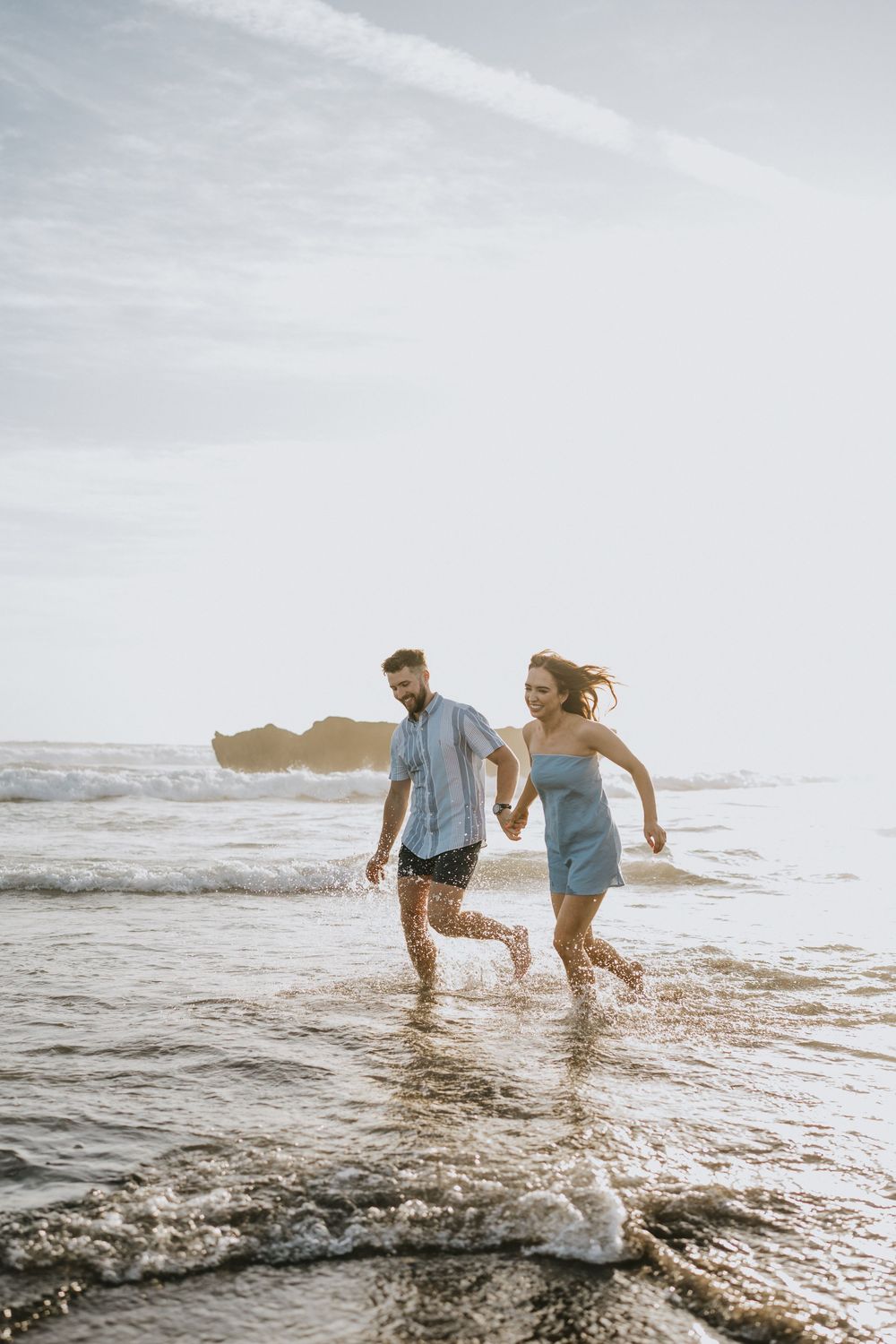 Two people running through shallow ocean water at the beach during golden hour.