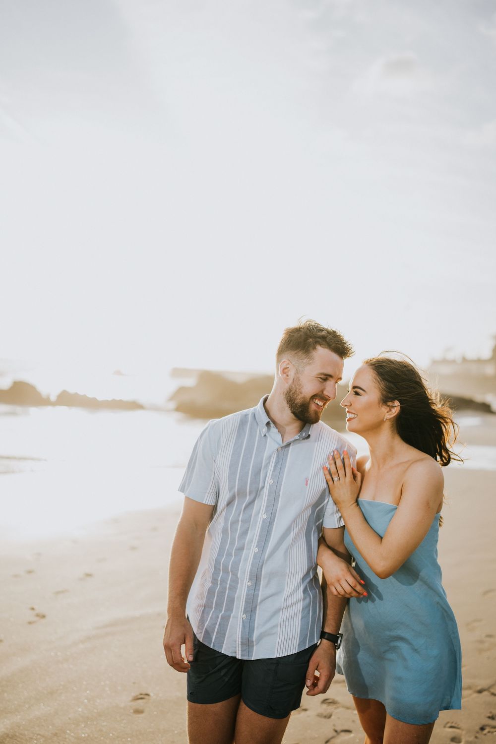 Couple sharing intimate moments on a sandy beach with rocks in the background.