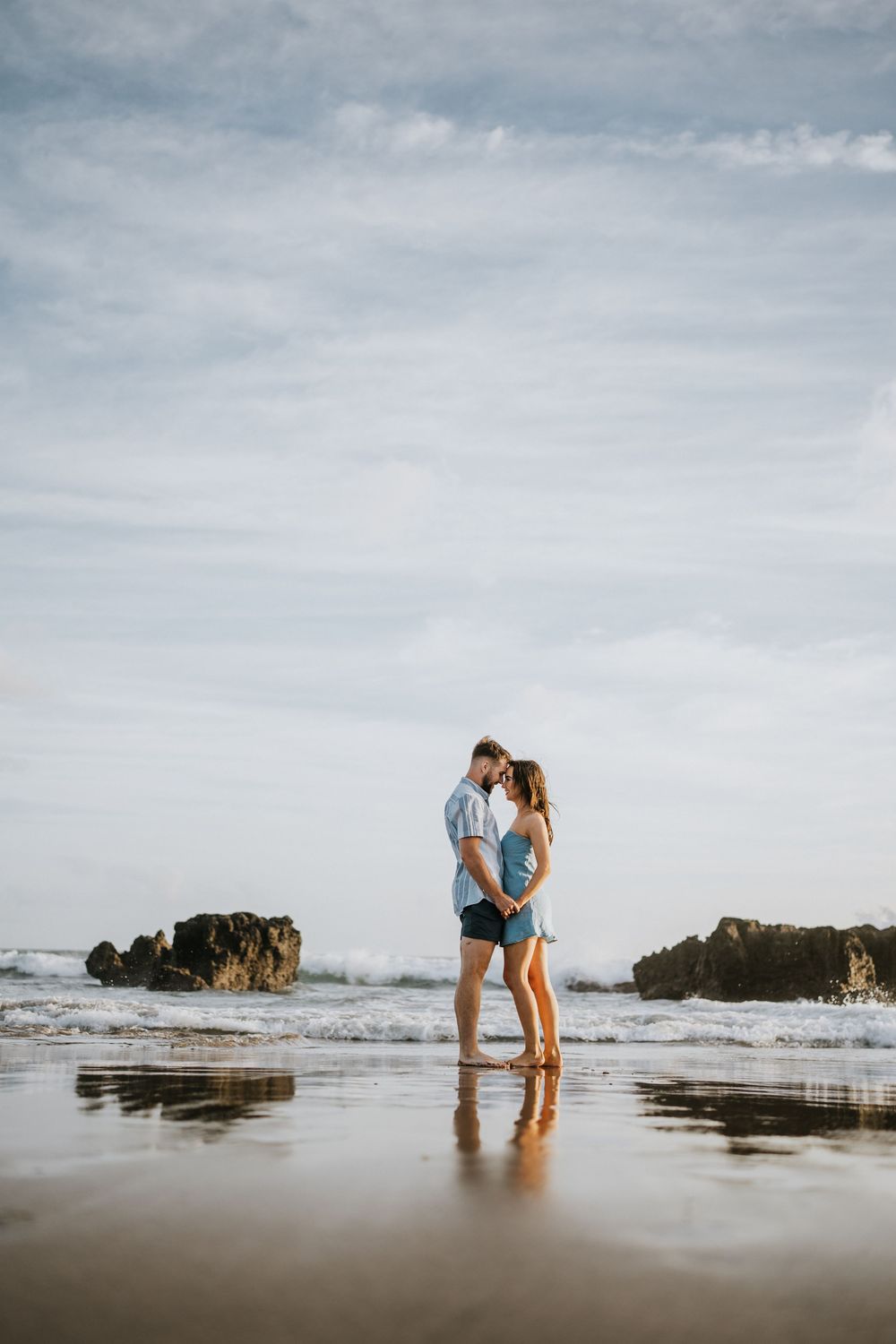 Couple standing together on wet sand with ocean waves and rocky formations in view.