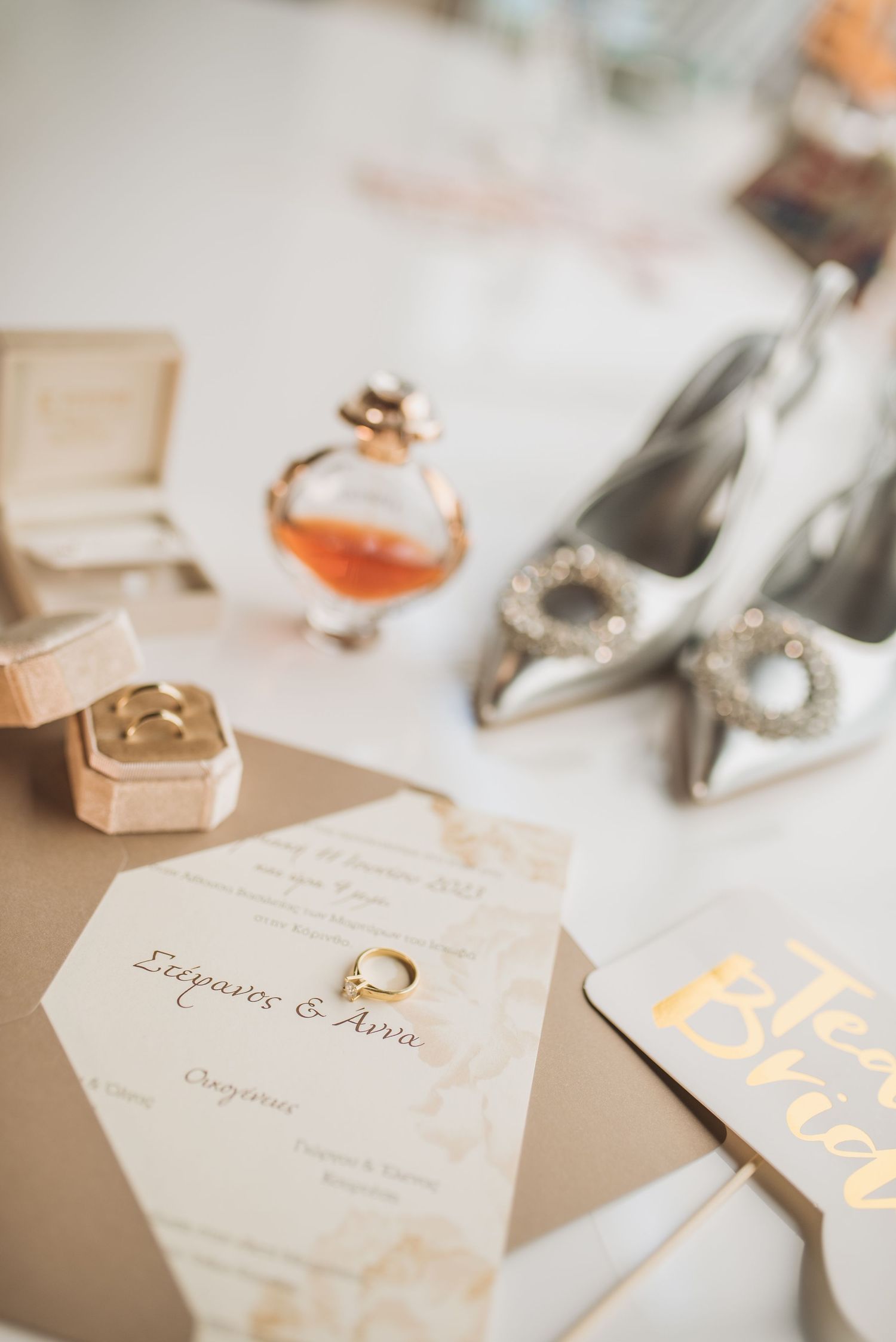 Elegant wedding accessories including perfume, silver heels with crystal details, rings, and invitation cards laid on white surface.