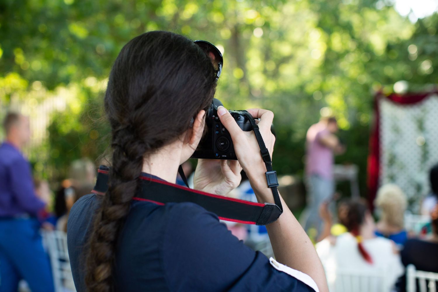 A crowd gathers outdoors at a social event under green trees and natural lighting.