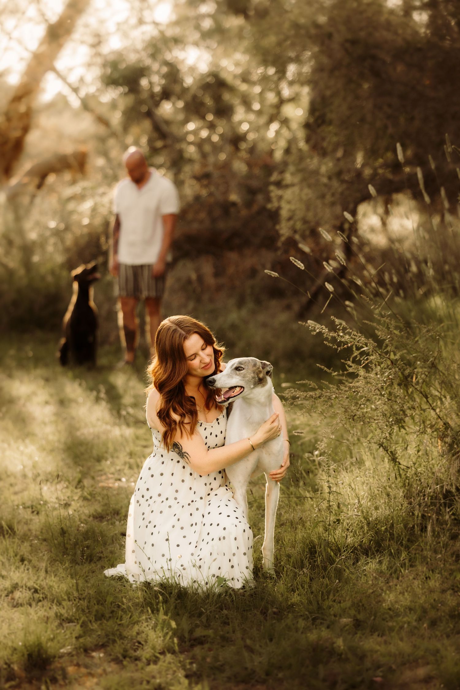 Woman in polka dot dress hugging a white dog outdoors, man with black dog visible in background among green trees.
