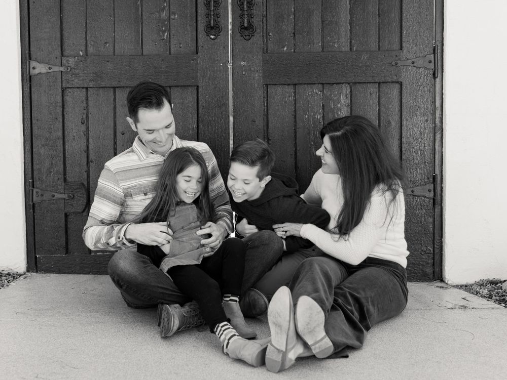 A family sits together on the ground reading a book against rustic wooden doors.