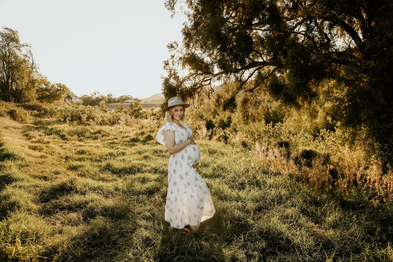 A person in a white flowing dress stands in a sunlit meadow surrounded by golden light and trees.