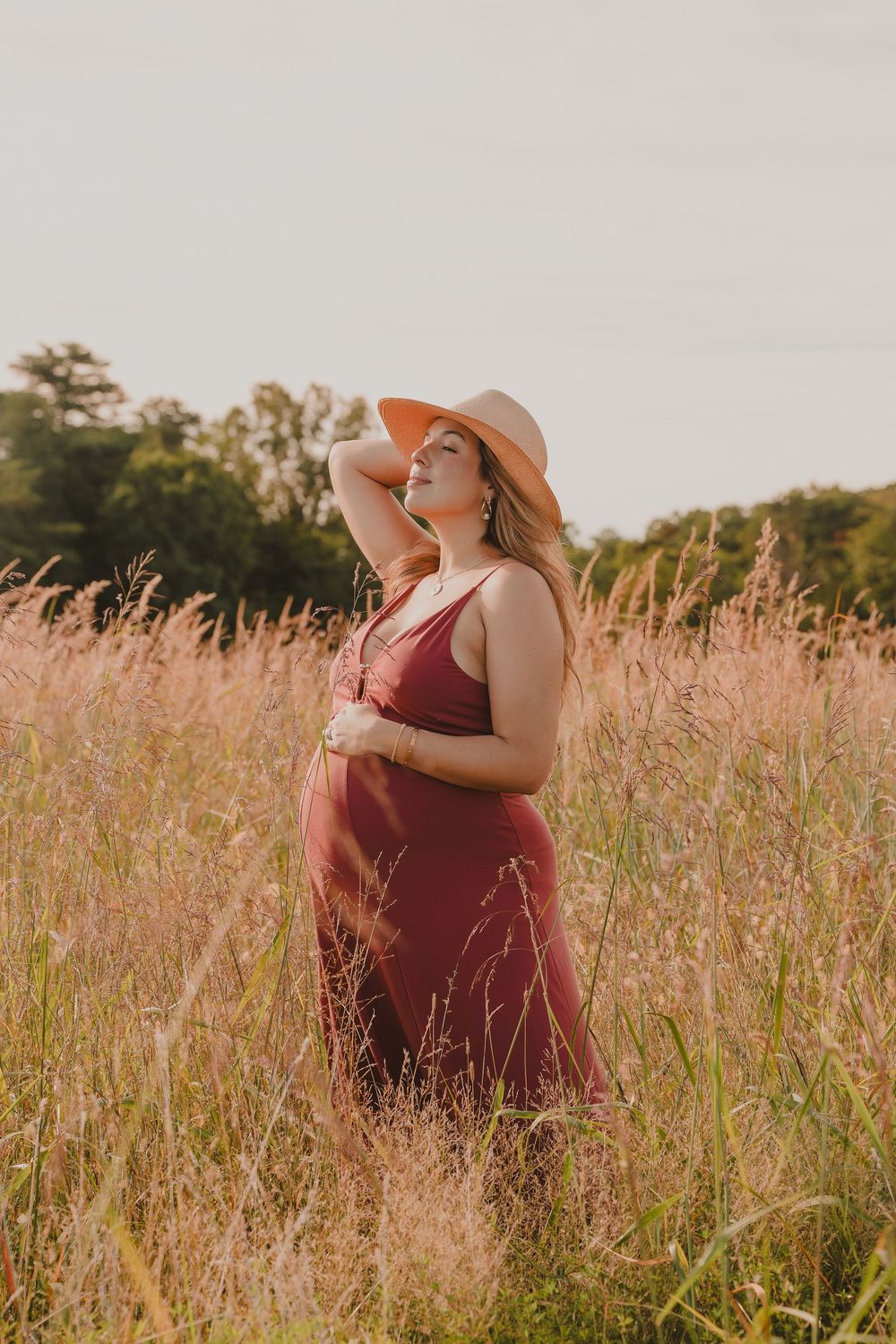 A dreamy sunset photoshoot in a field of tall grass with a person in a burgundy dress and straw hat.