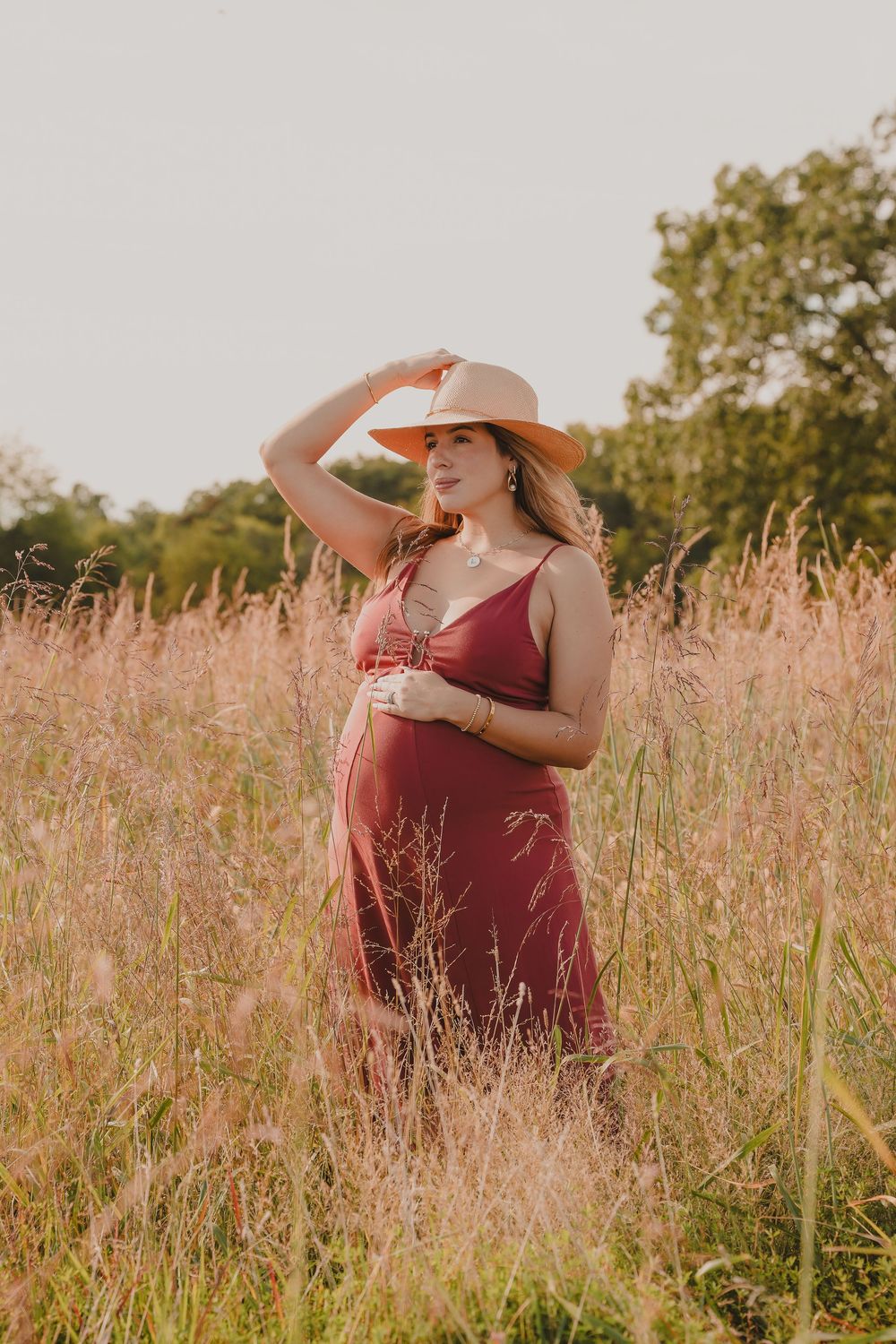A person wearing a burgundy dress and wide-brimmed hat poses gracefully in a field of tall wildflowers at sunset.
