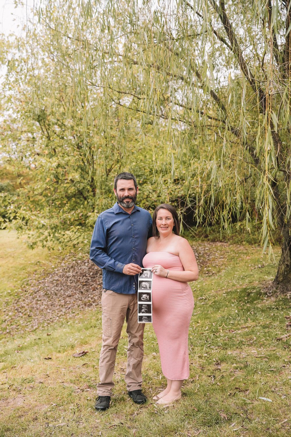 A couple stands outdoors in nature holding an ultrasound photo, sharing their pregnancy announcement in autumn scenery. Leesburg, Virginia
