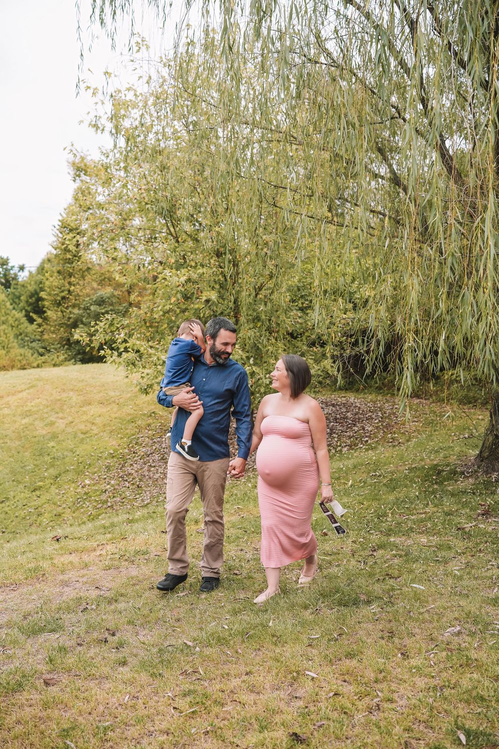 A couple in casual attire shares a playful moment on a grassy hillside with willow trees in the background. Leesburg, Virginia