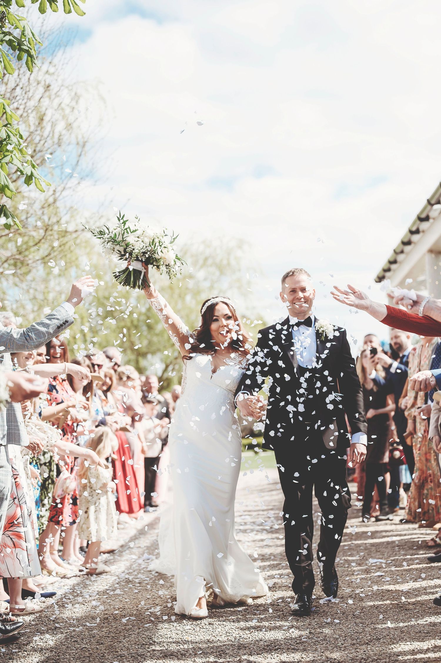 Wedding guests showering newlyweds with white confetti while celebrating outdoors beneath trees on a sunny day.