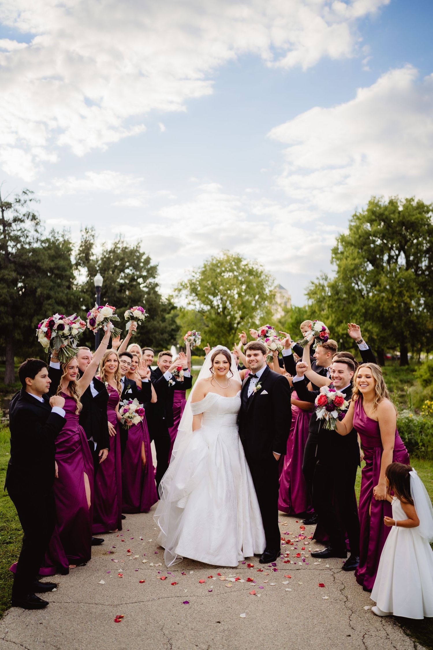 Wedding party celebrating outdoors with flower petals being tossed in the air as guests cheer in burgundy attire.