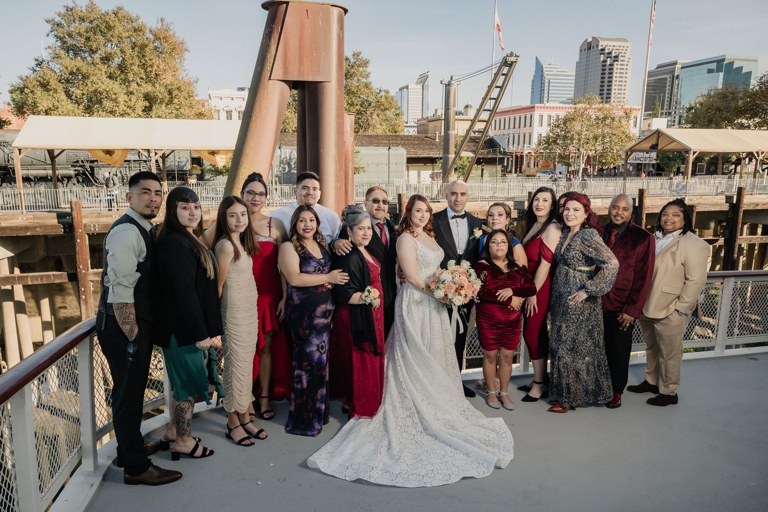 A wedding party group poses on a deck overlooking a marina with city buildings visible in the background at sunset for Sacramento wedding.