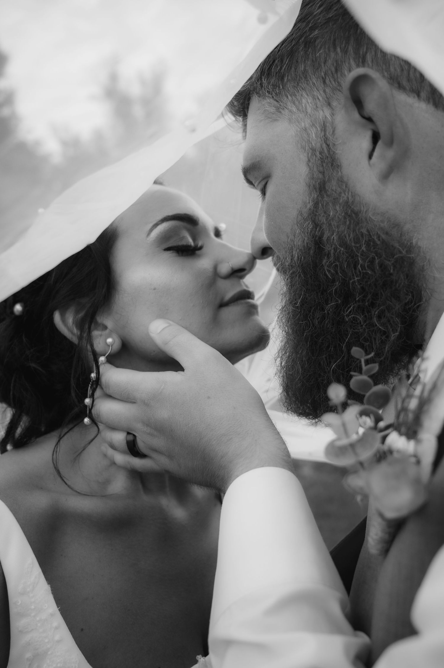 Black and white photo of bearded groom tenderly holding brides face for a kiss under veil.