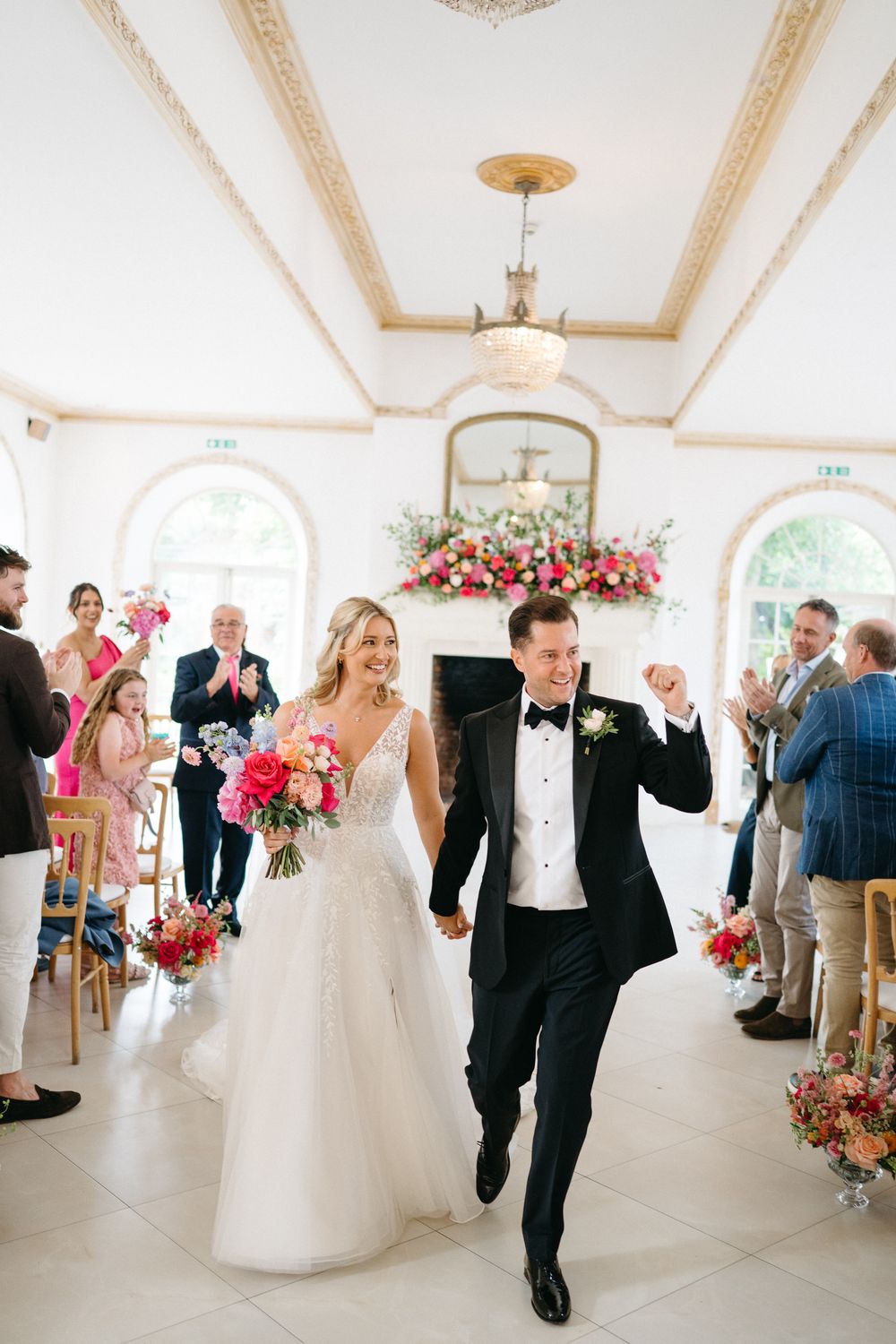 Wedding celebration scene in a bright ballroom with floral arrangements and a crowd of guests.