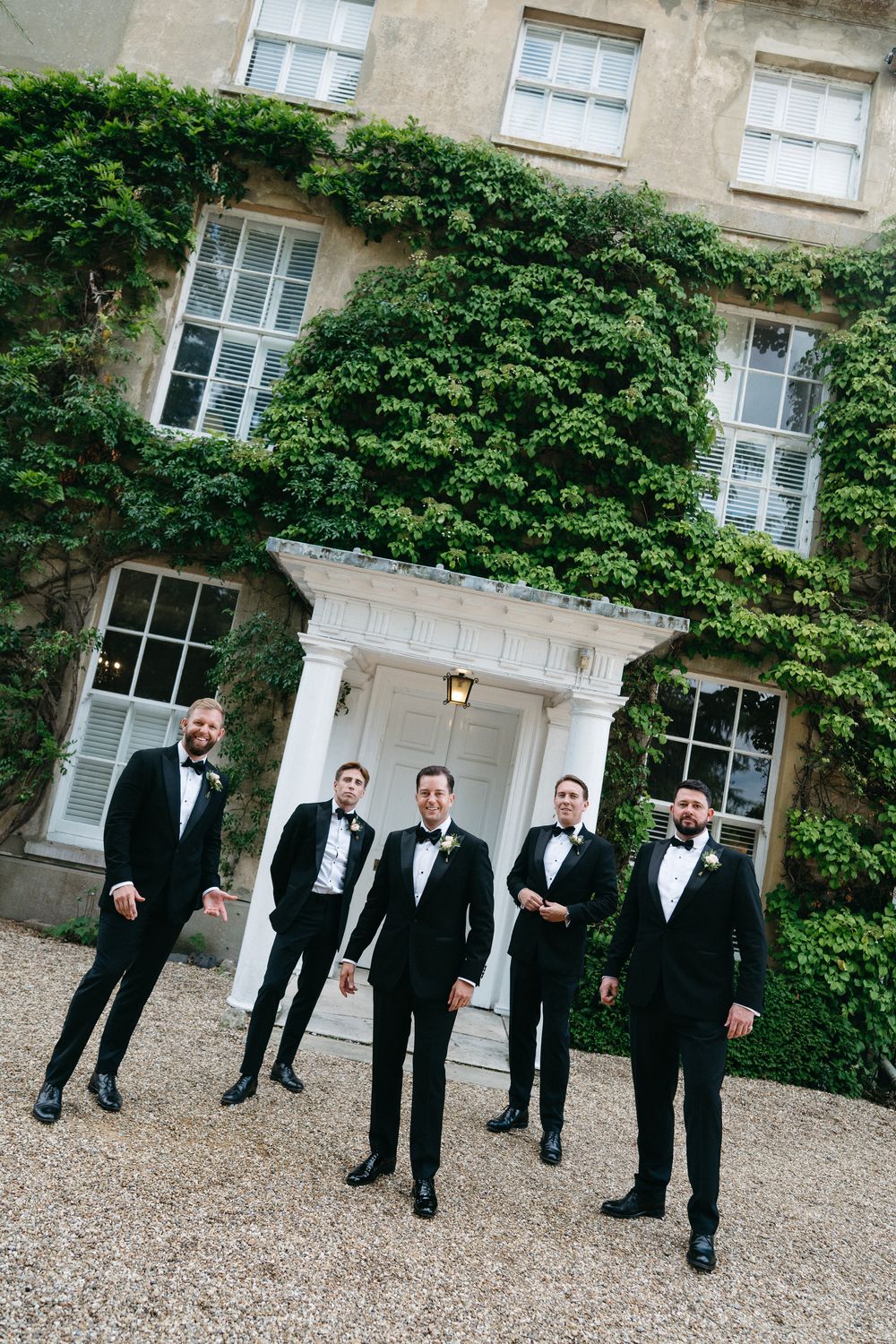 Groomsmen in black tuxedos stand in diagonal line formation on gravel path outside ivy-covered mansion wall.