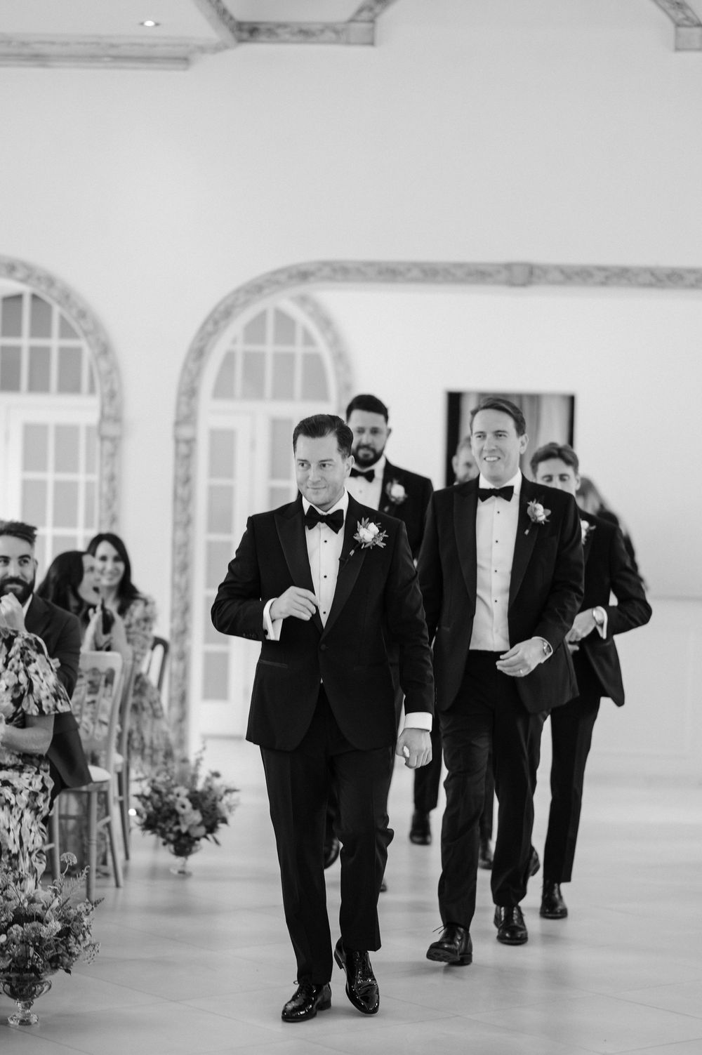 Groomsmen in black tuxedos line up playfully during a wedding celebration in a bright white room.
