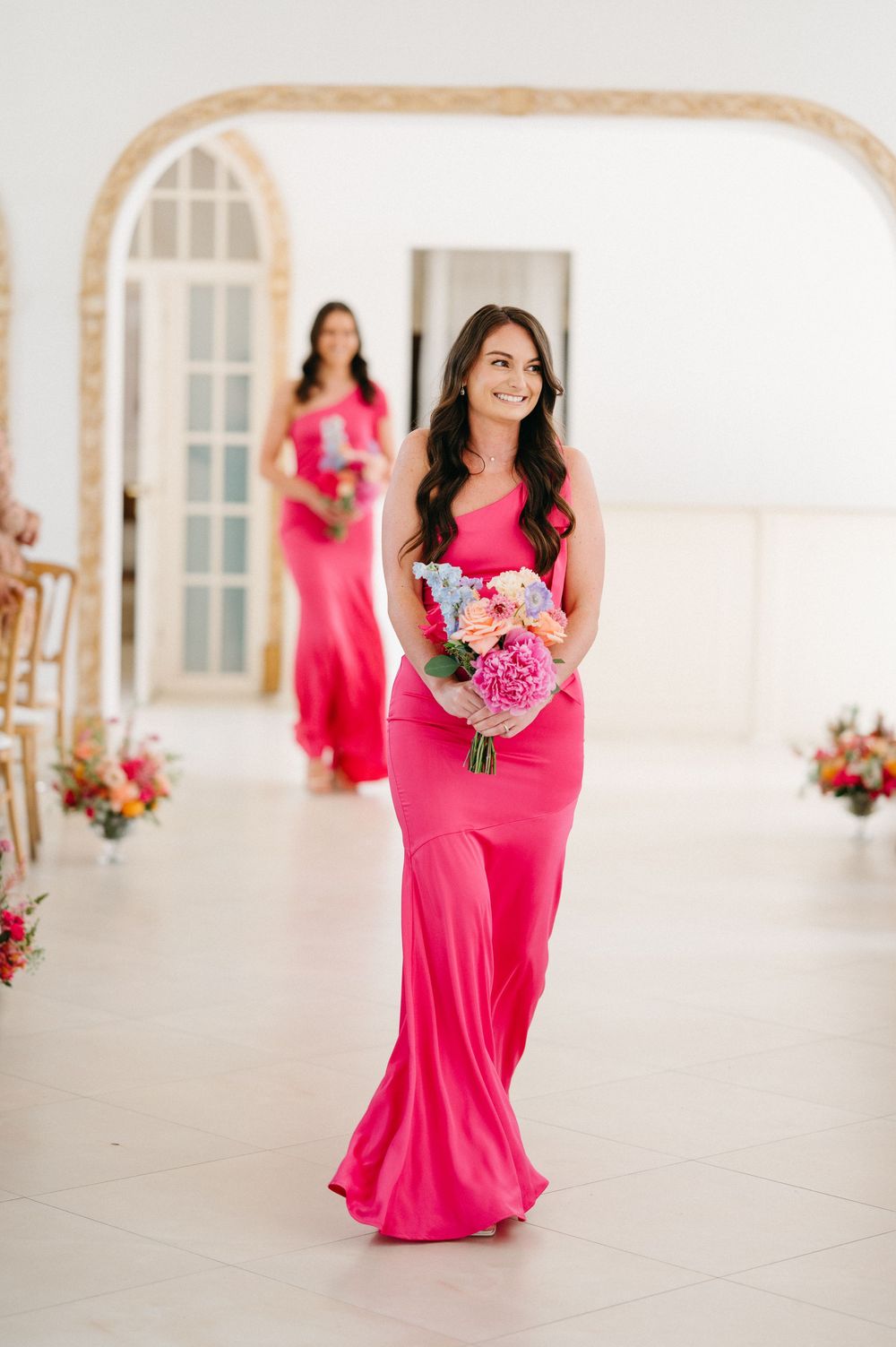 Bridesmaids in bright pink floor-length dresses carrying floral bouquets in an elegant white venue.
