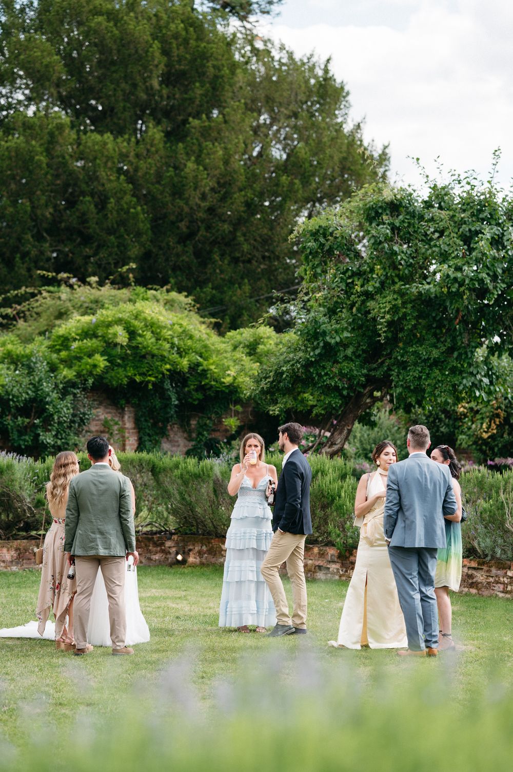 Wedding party poses in a garden setting with lush greenery and stone walls.