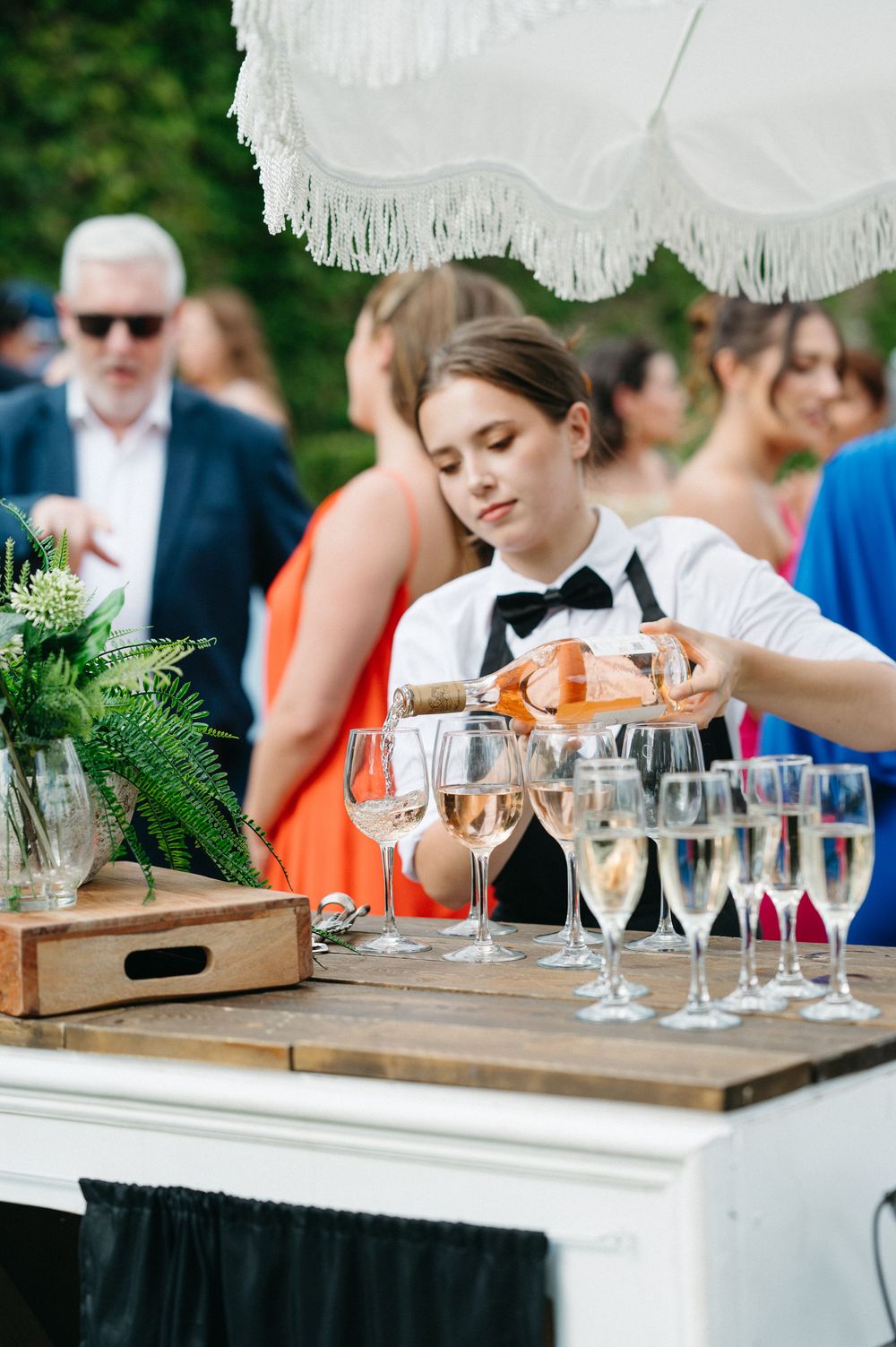 Crystal wine glasses lined up on wooden serving trays at an outdoor event with white parasol decorations.
