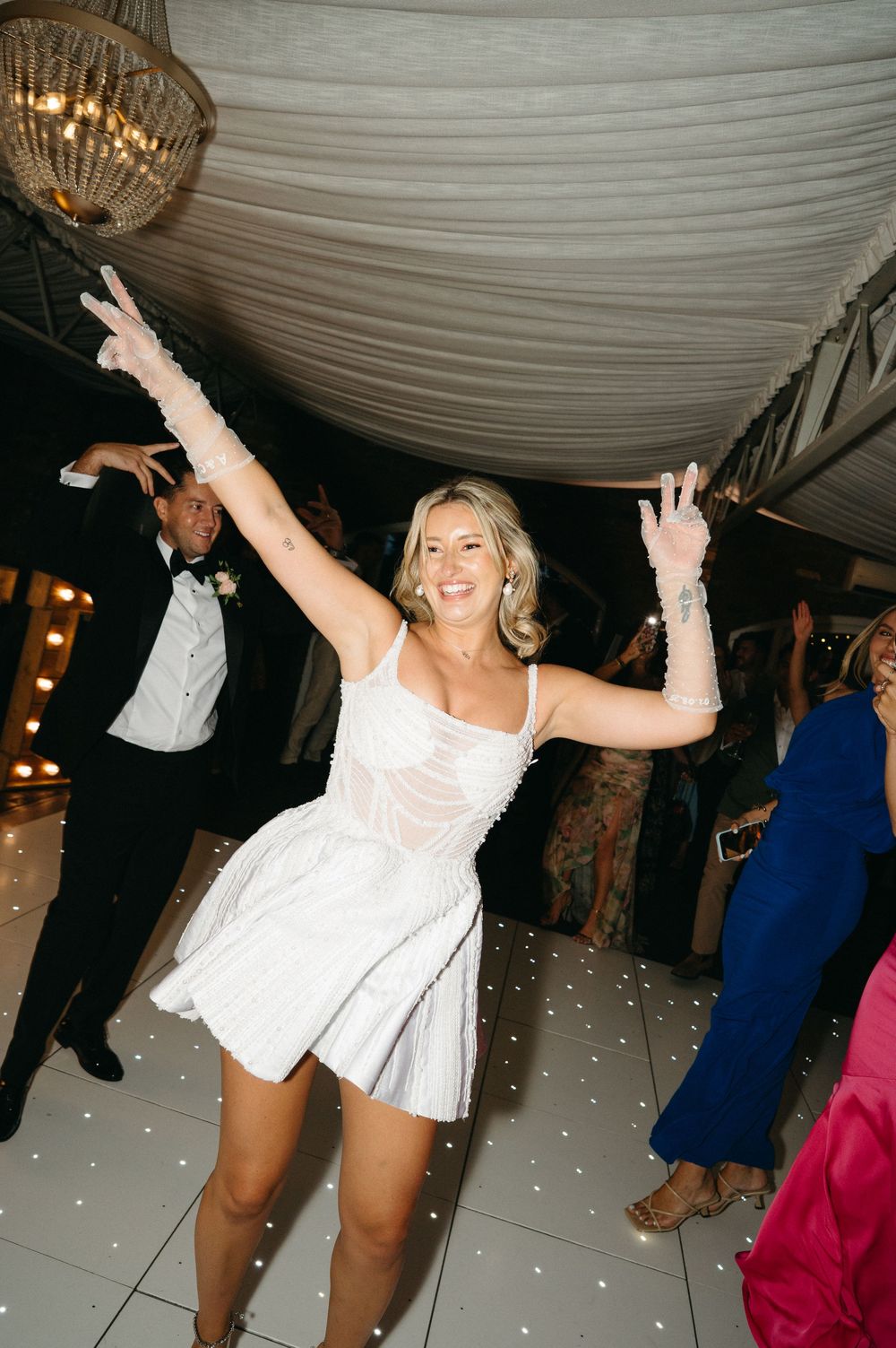 People dancing and celebrating on an illuminated white dance floor under a draped tent ceiling with crystal chandeliers.