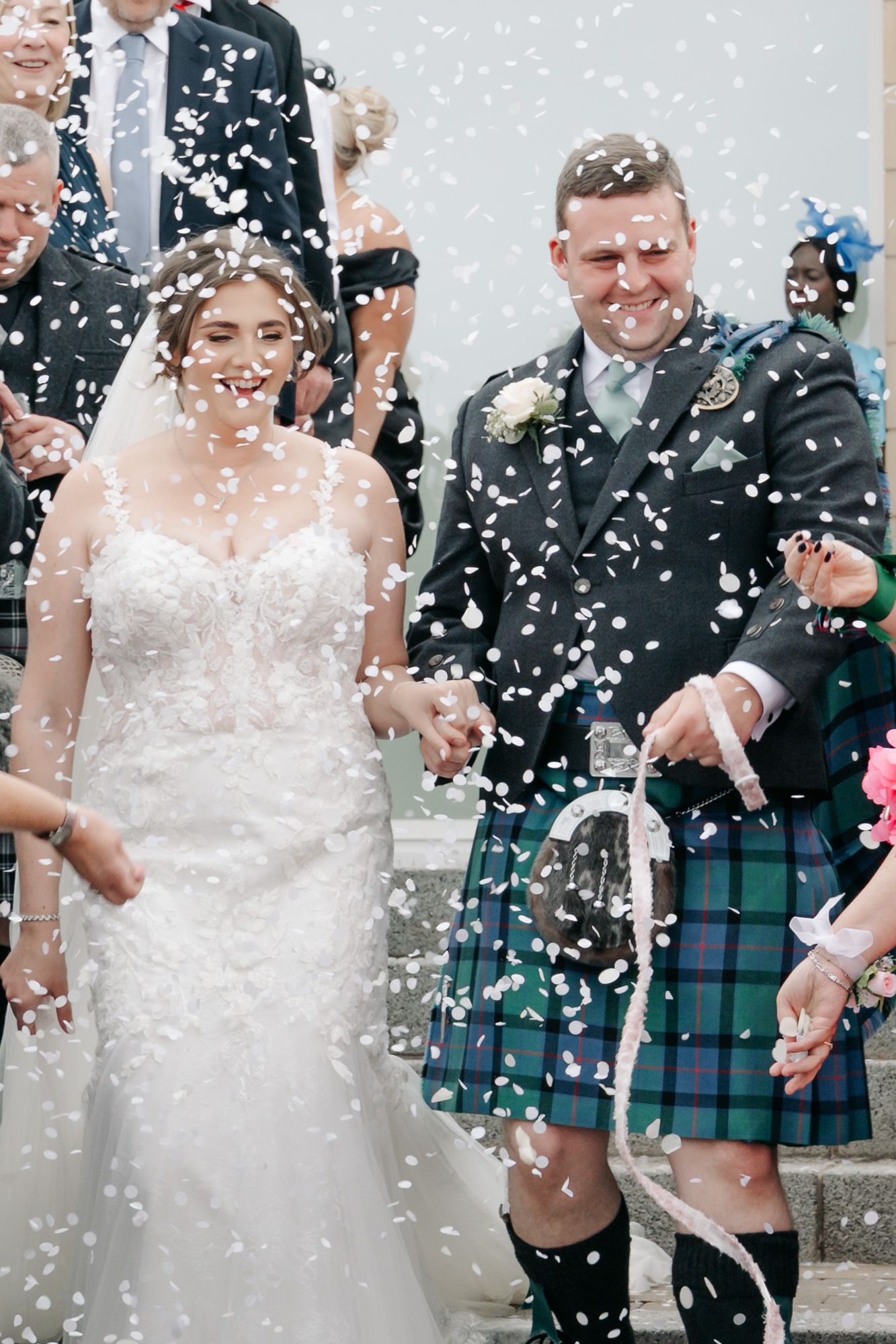 Joyful newlyweds surrounded by falling confetti, with the groom wearing a traditional tartan kilt.