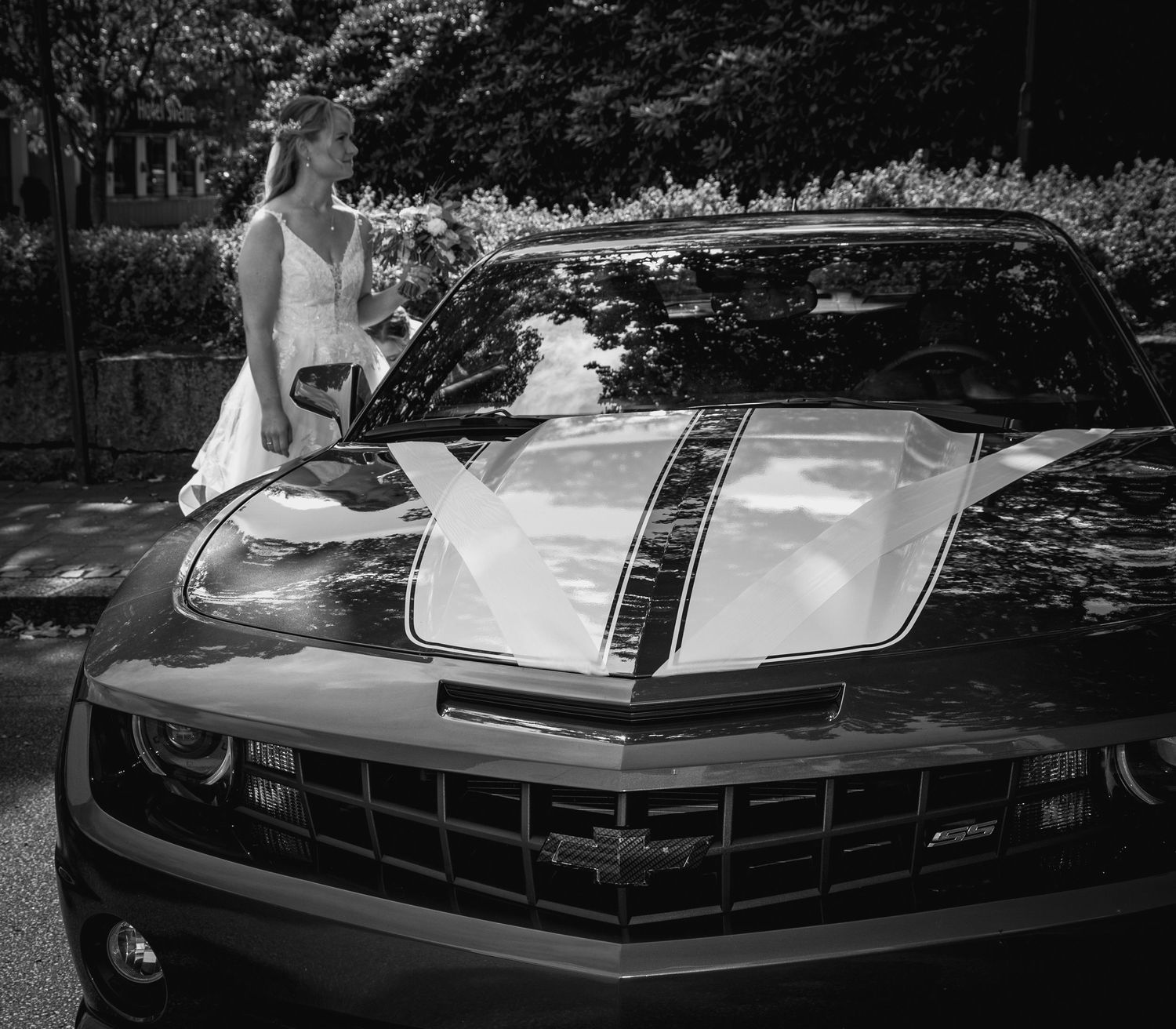 Elegant bride in white gown poses beside classic Chevrolet Camaro in black and white photo.
