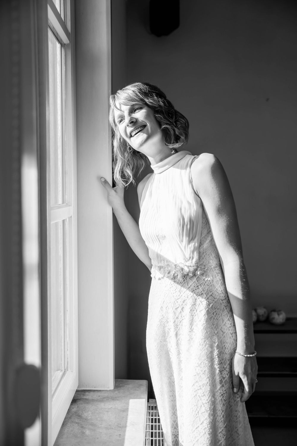 A joyful person in a white lace dress stands near a window basking in natural light.