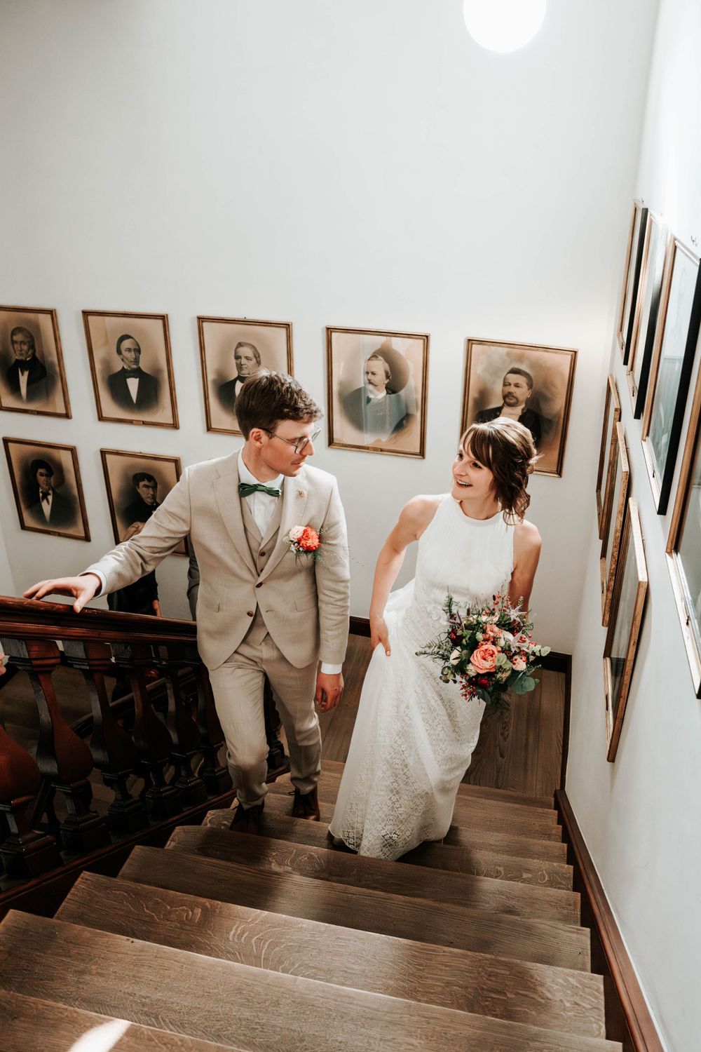 A joyful couple in wedding attire walk down wooden stairs together, with vintage portrait photographs lining the wall.