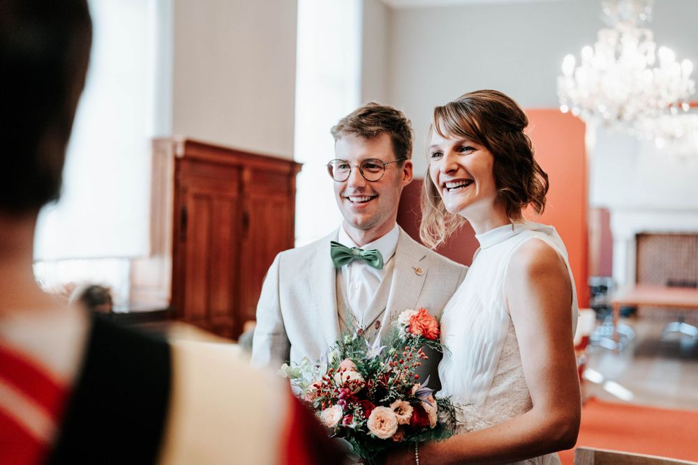 Happy couple shares joyful laughter during wedding ceremony in elegant room with chandelier and floral bouquet.