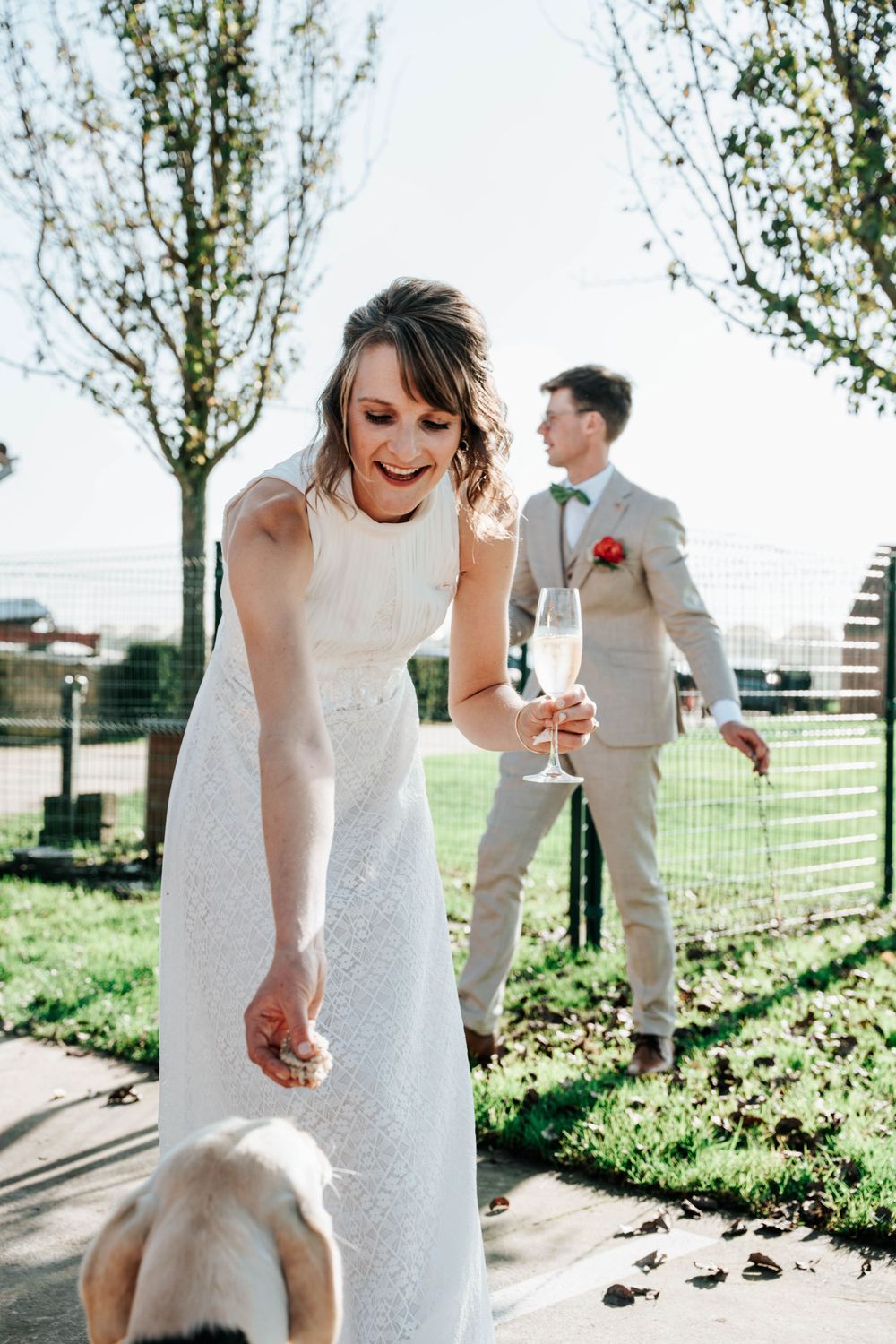 Wedding couple in outdoor garden setting with white dress and light gray suit enjoying champagne on sunny day.