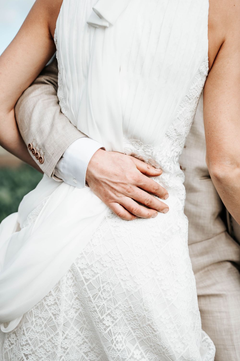 Close-up of hands embracing at waist in white lace wedding dress and beige suit.