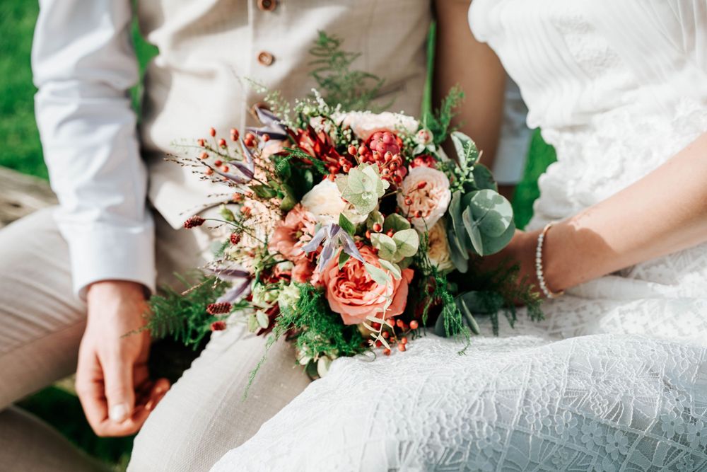 Beautiful peach and coral wedding bouquet with roses and eucalyptus held between two people in white and beige attire.