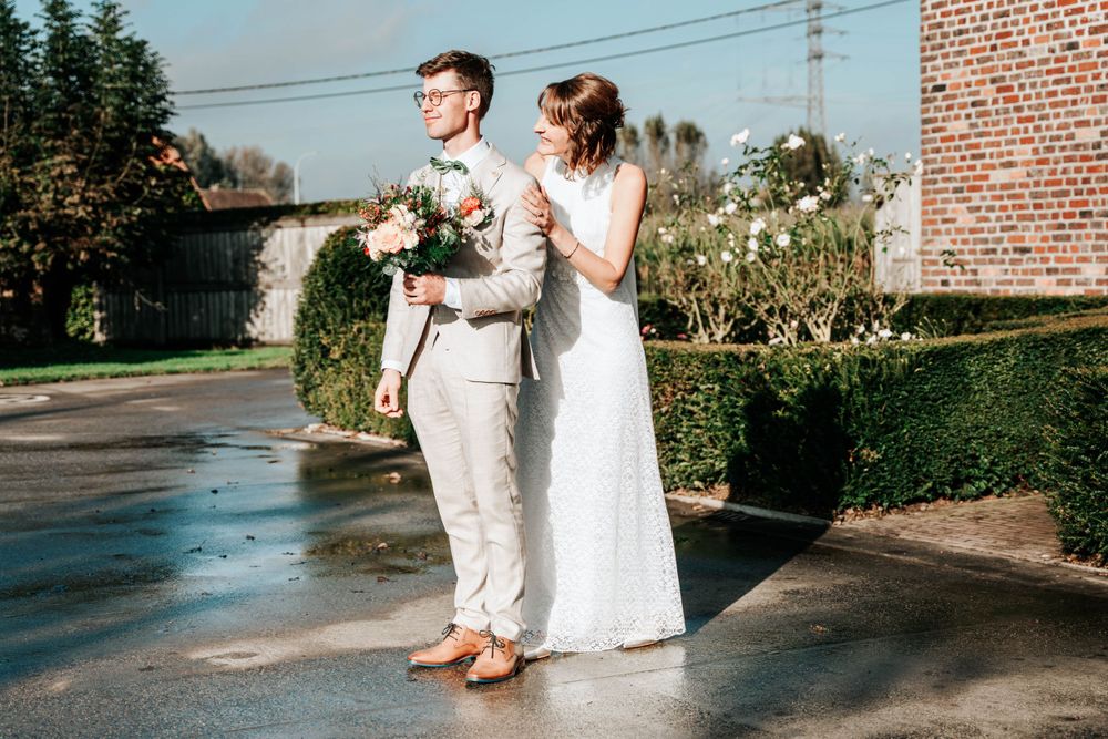 A couple stands together outdoors during a wedding celebration, the bride in white lace and groom in light beige.