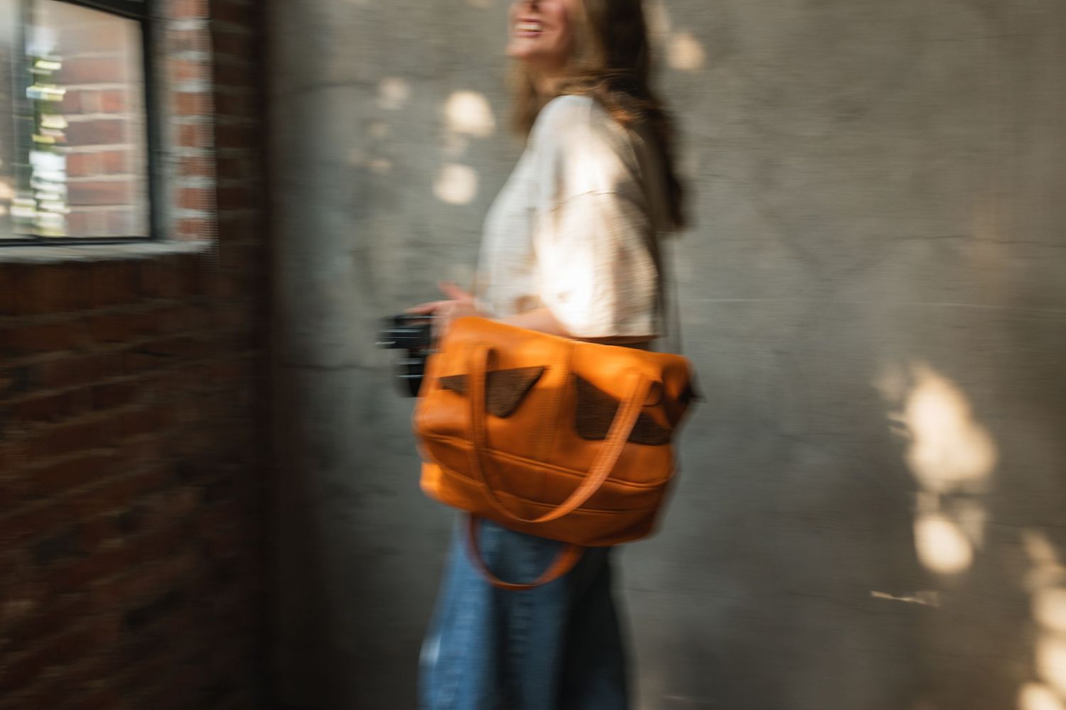 Person in blue jeans and brown jacket walks down urban street carrying orange leather bag.