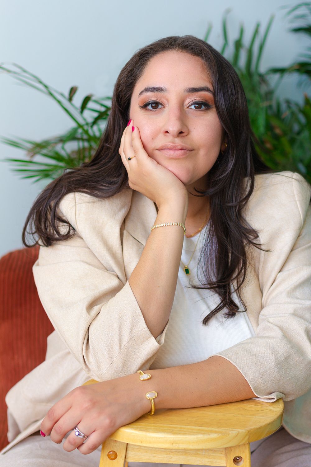 Professional headshot of person in cream blazer resting chin on hand with plants in background.