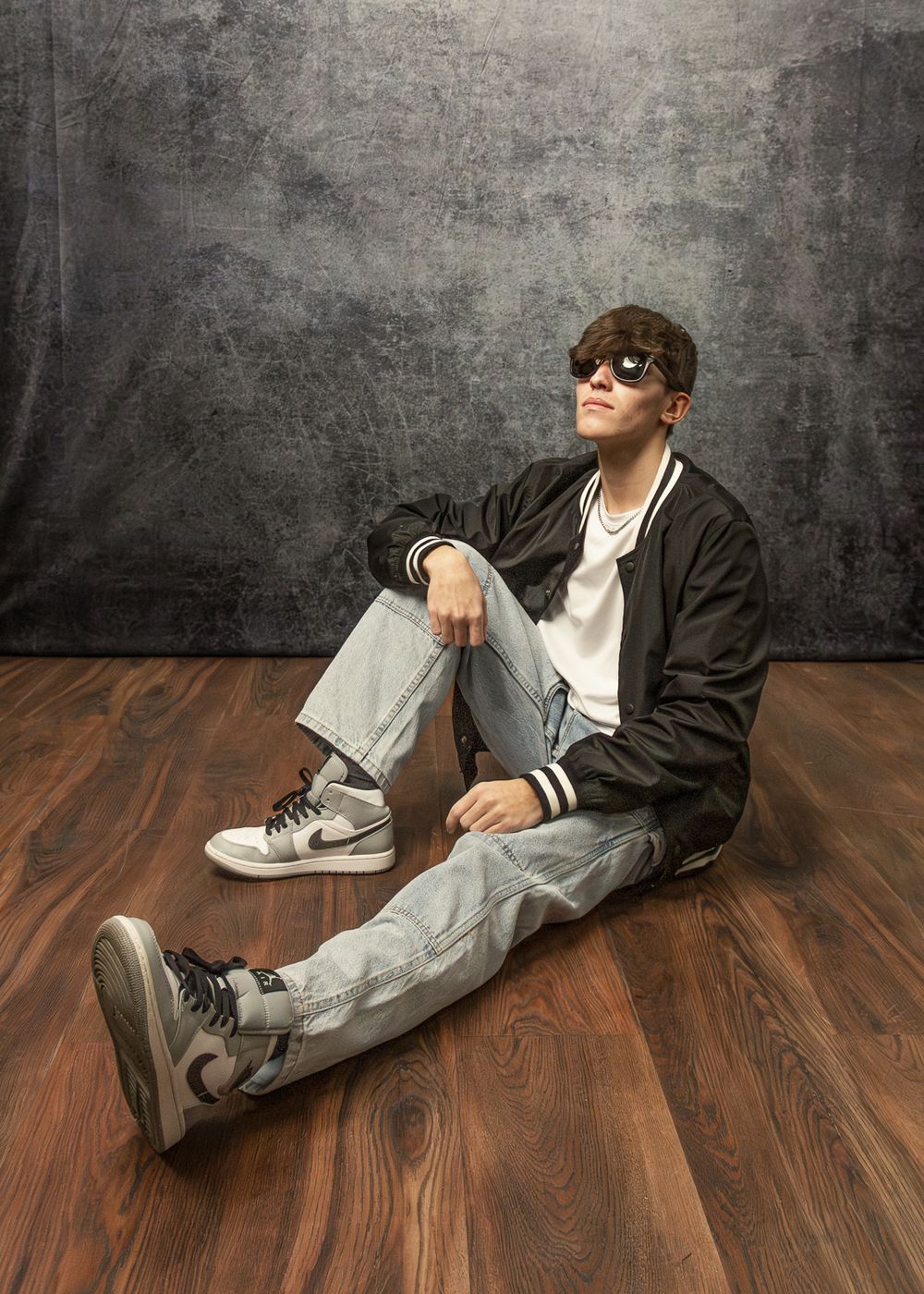 Casual portrait of person in sunglasses and jacket sitting on wooden floor against grey backdrop.