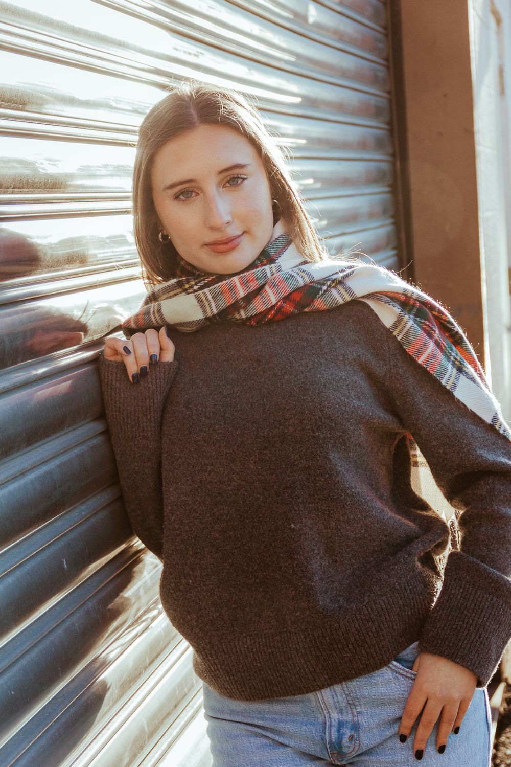 Fashion portrait against metal door wearing plaid scarf and brown sweater in natural light.