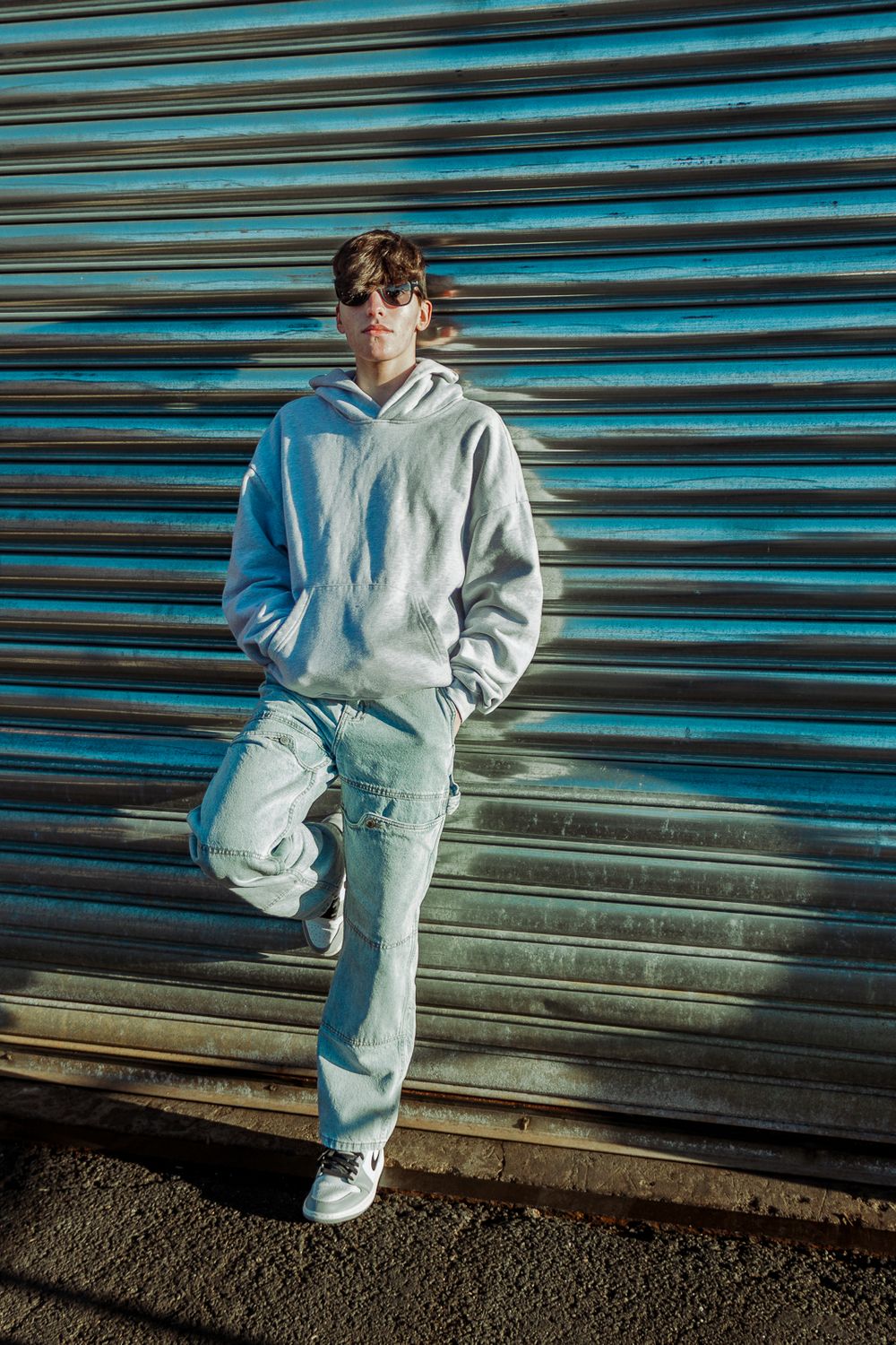 Person in casual light-colored outfit posing against blue corrugated metal wall in urban setting.