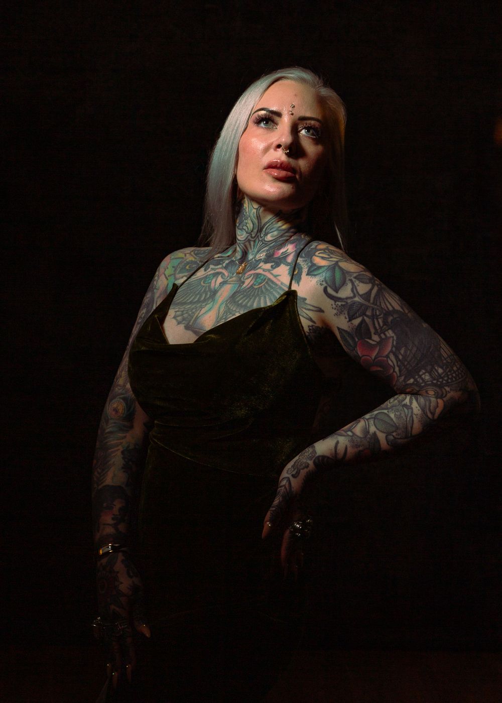 Figure in black dress with colorful tattoo sleeves poses dramatically against dark background.