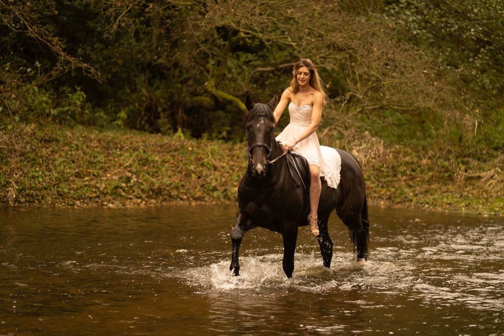 A dreamy portrait of a rider in white dress crossing a stream on a black horse.
