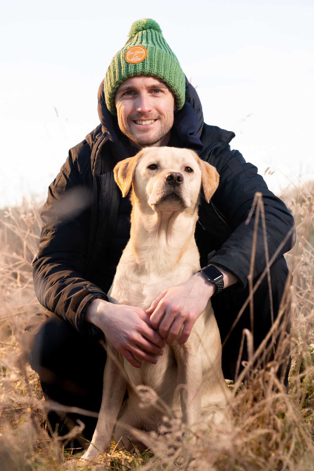 A yellow Labrador retriever poses with someone wearing a green knit hat and black jacket in autumn field.