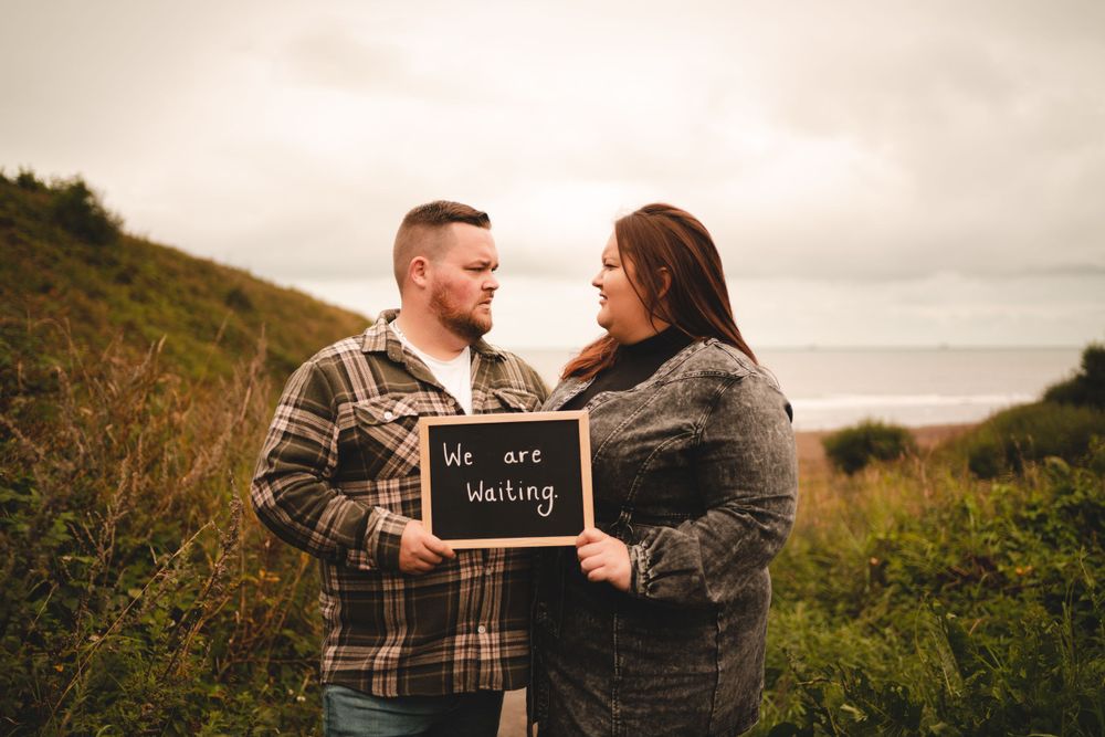 A couple holds a chalkboard sign announcing their engagement on a cloudy hillside.