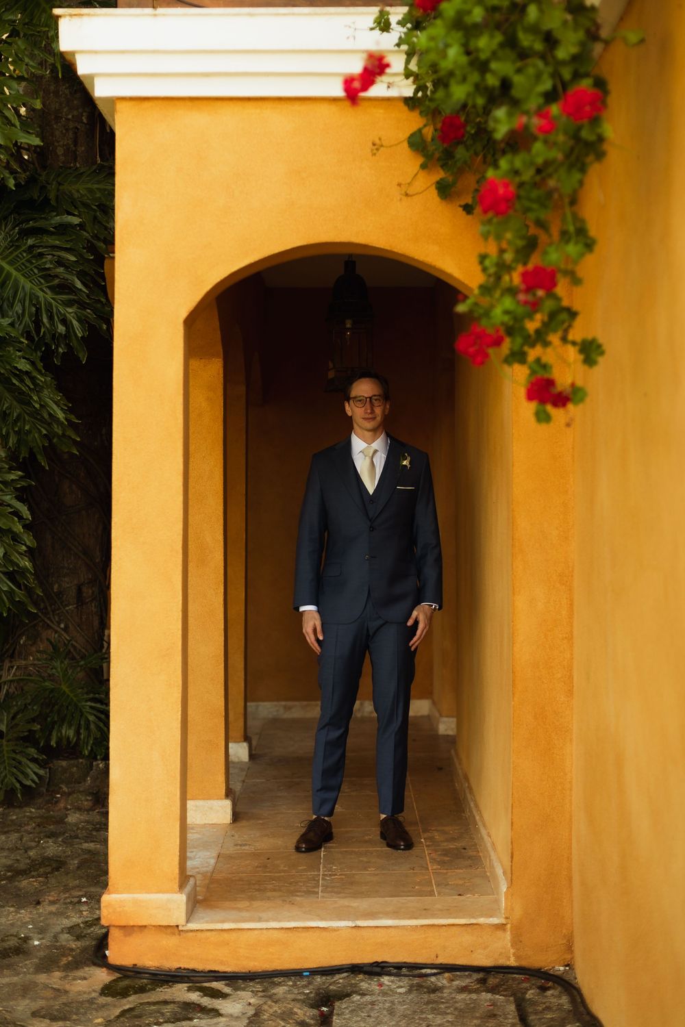 A person in a navy suit stands in an arched yellow stucco doorway with red flowers hanging above.