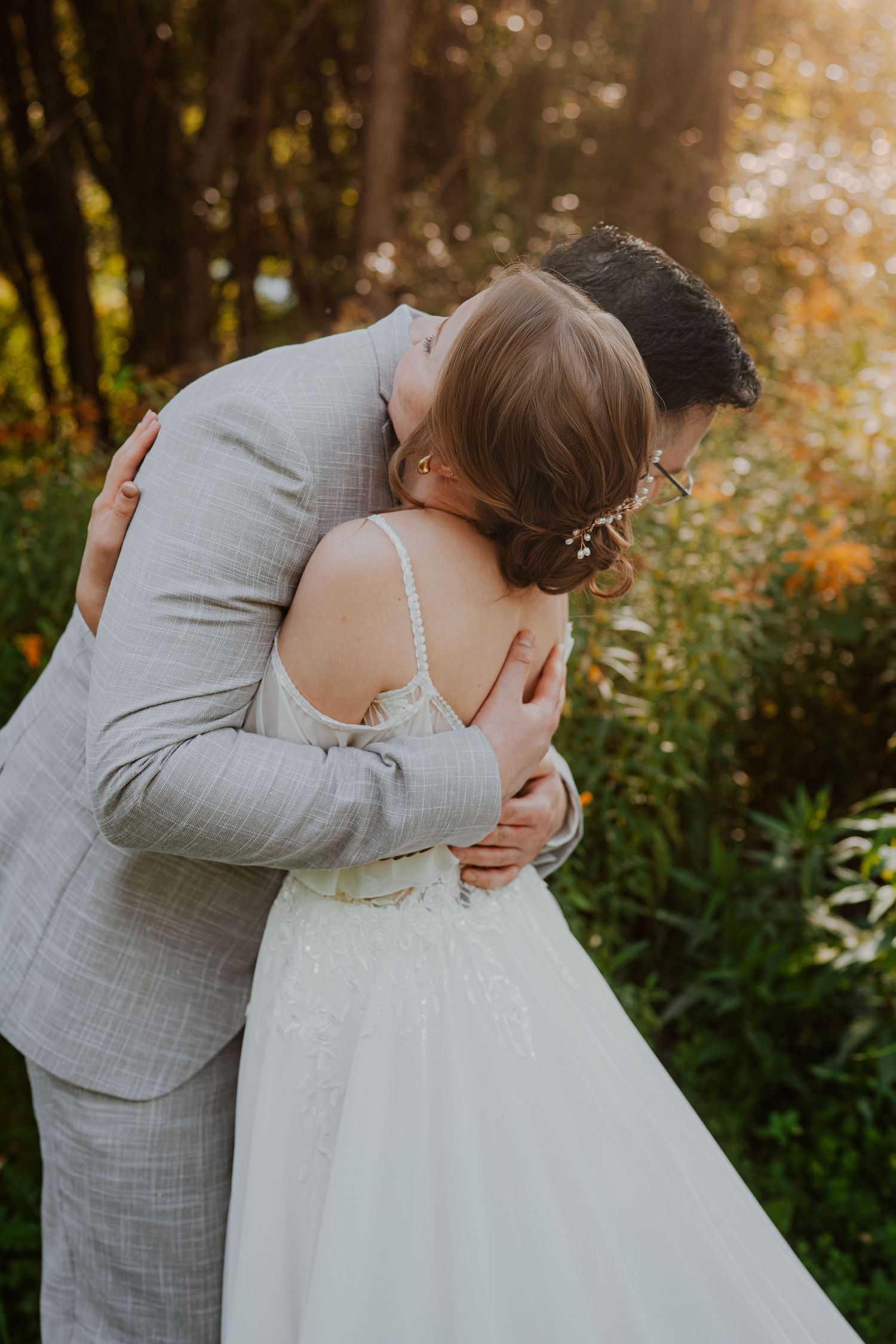 A romantic couple in white clothing shares an intimate moment in a wooded setting during golden hour.