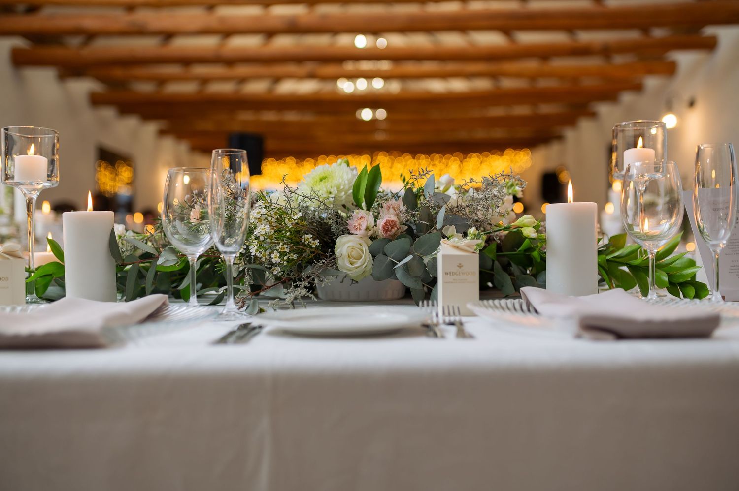 Elegant wedding table setting with white linens, wine glasses, and fresh greenery centerpiece under wooden beamed ceiling.