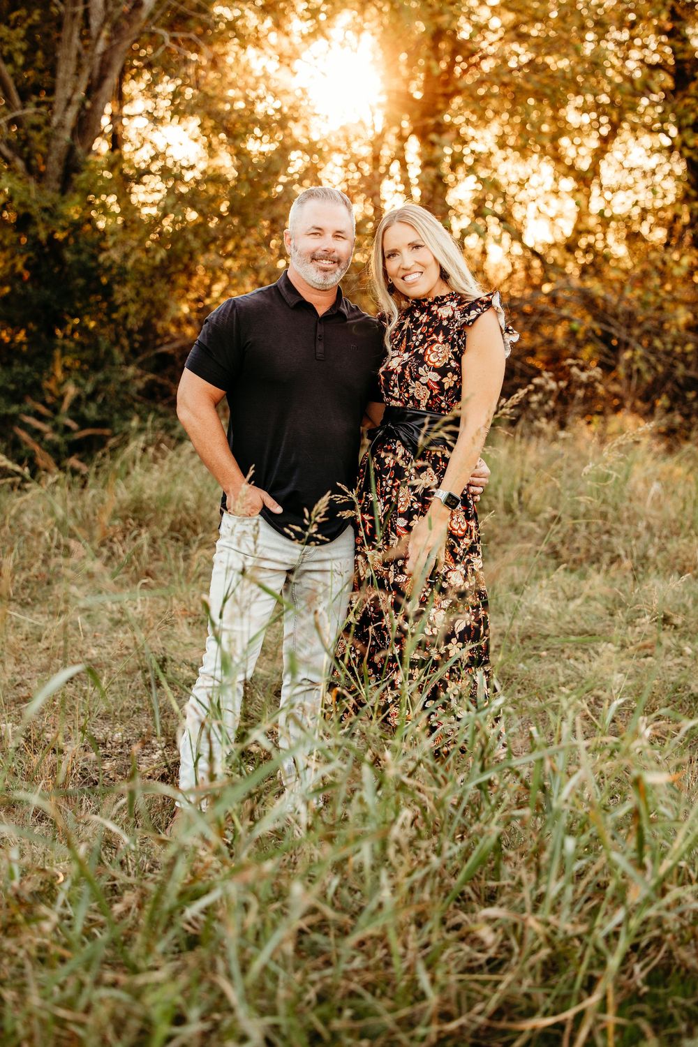 A couple poses together in a grassy field at sunset with golden sunlight filtering through autumn trees behind them.
