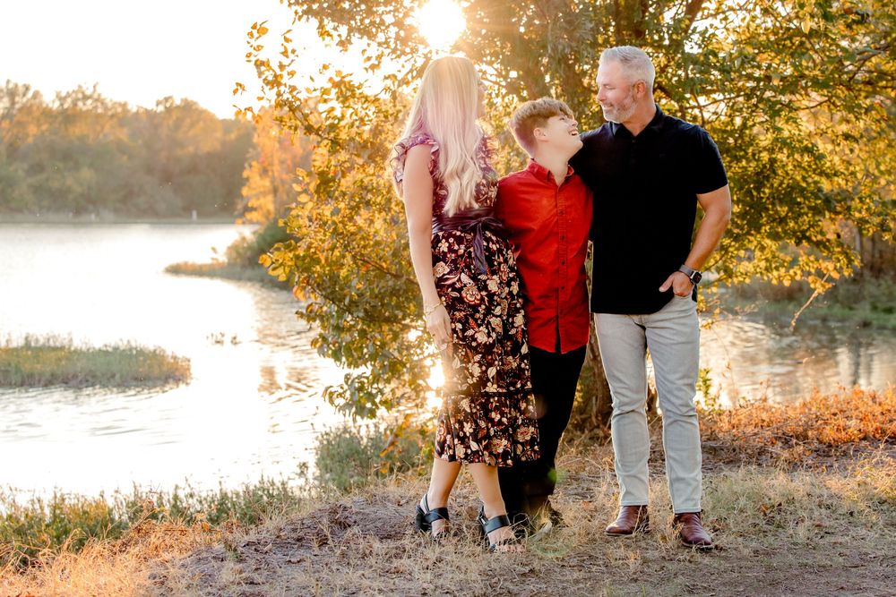 Family walking along a lakeside path during golden hour sunset with autumn trees and sunlight glowing in background.