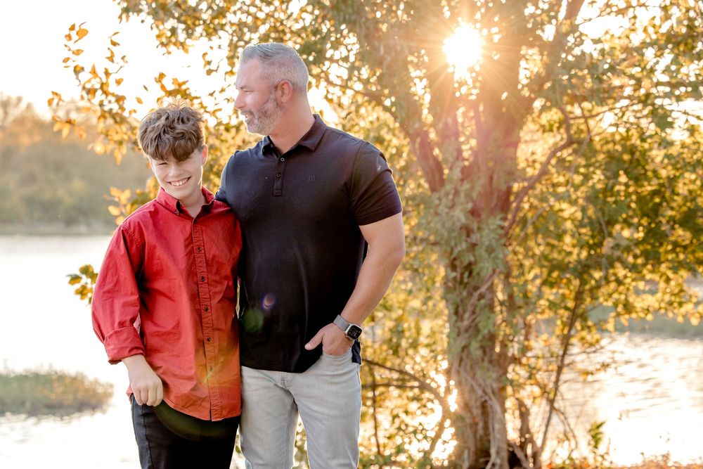 A parent and child share a tender moment outdoors during golden hour sunset with autumn trees in the background.