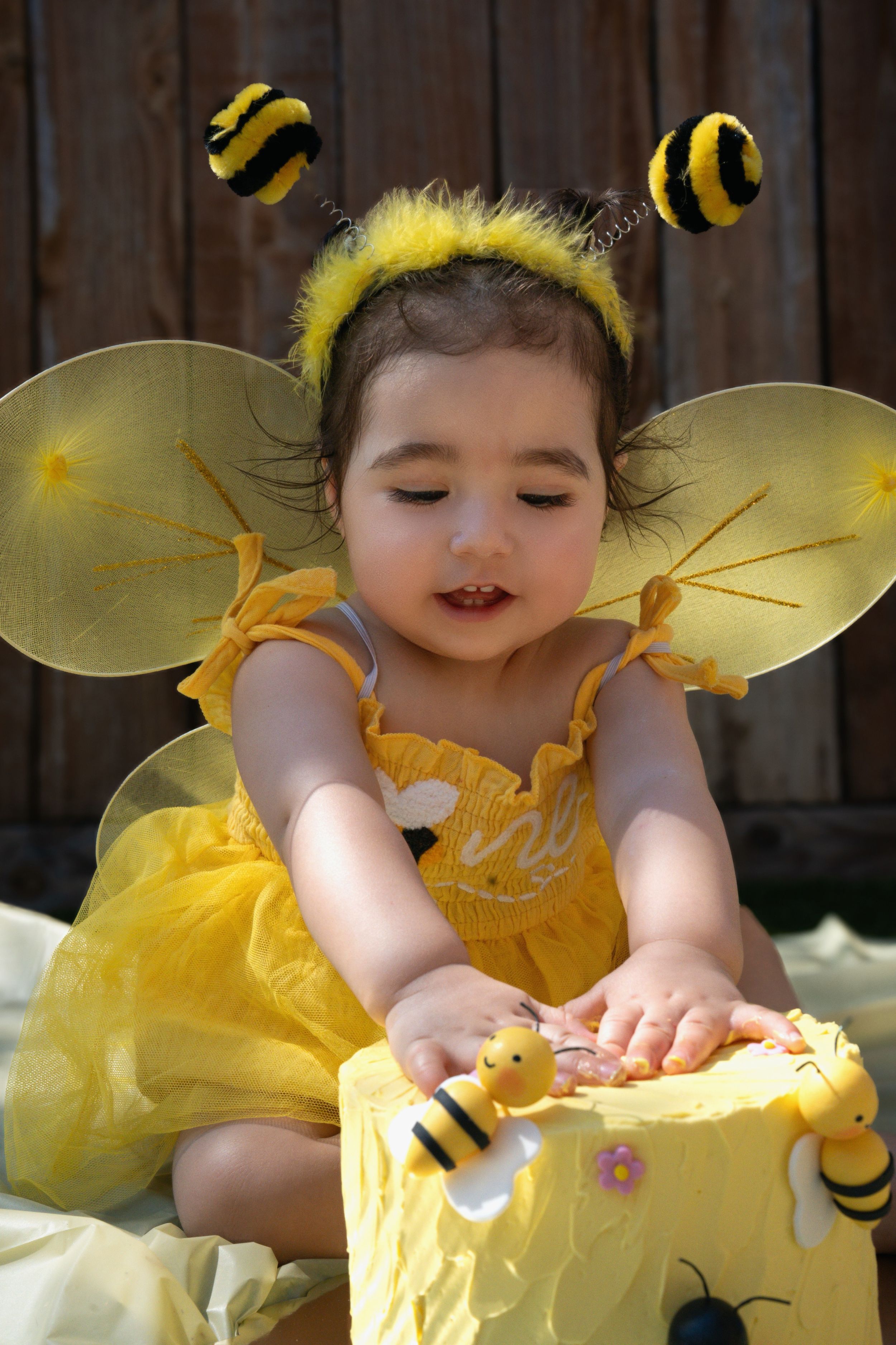 Baby in yellow bee costume exploring birthday cake during cake smash photoshoot.
