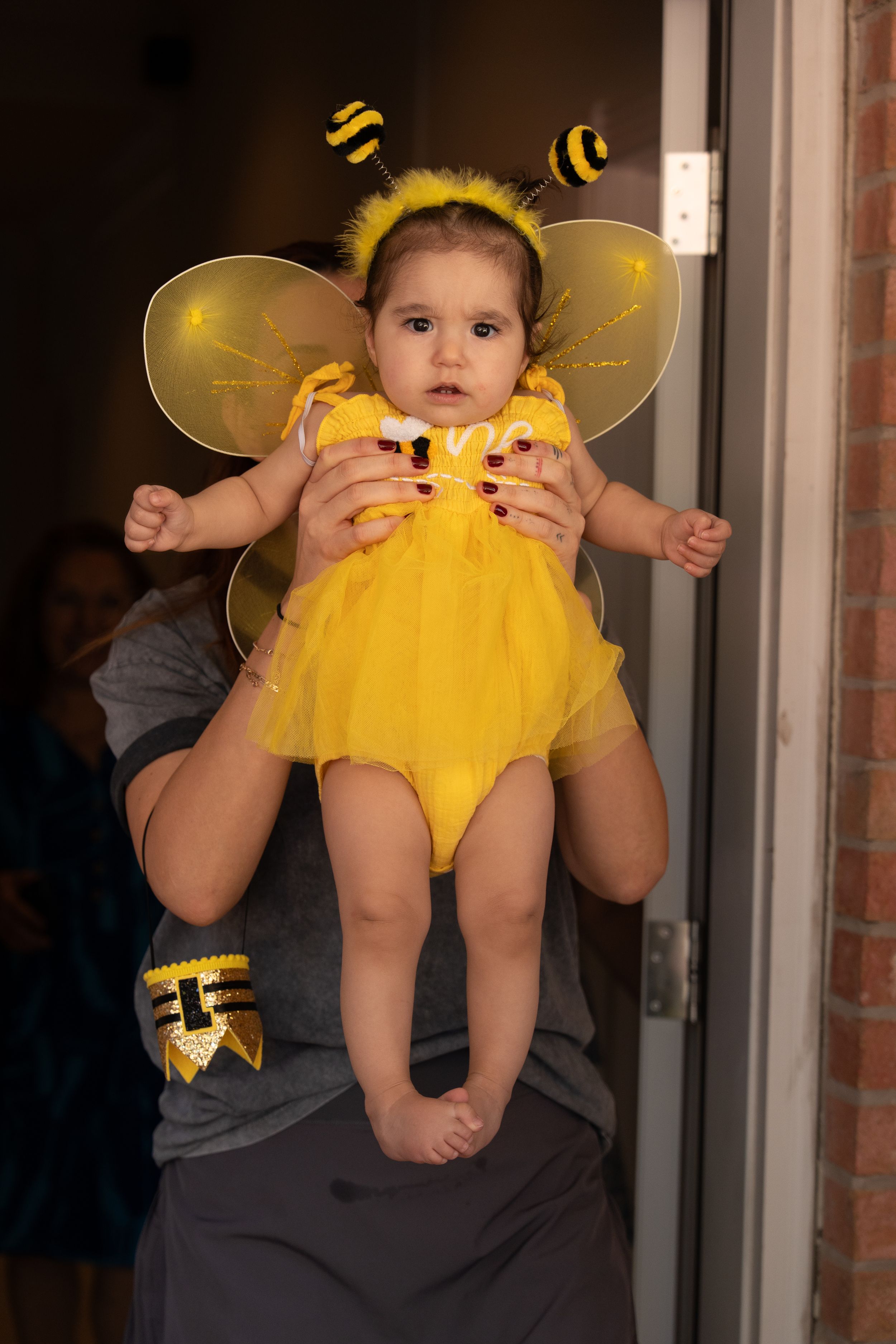 A small child dressed in a bright yellow bee costume being held up against a doorway.