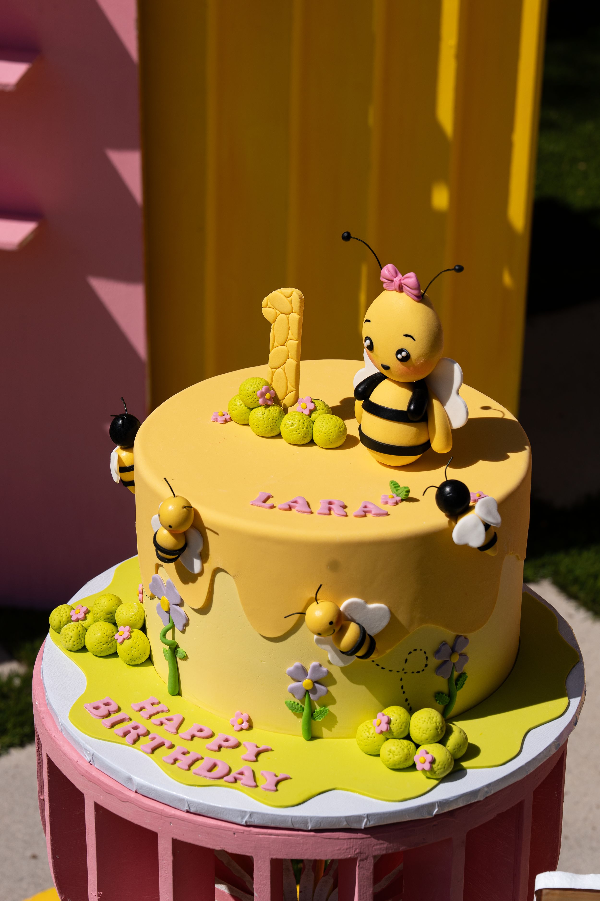A yellow fondant cake decorated with a cute bumble bee theme featuring small green accents and pink flowers.